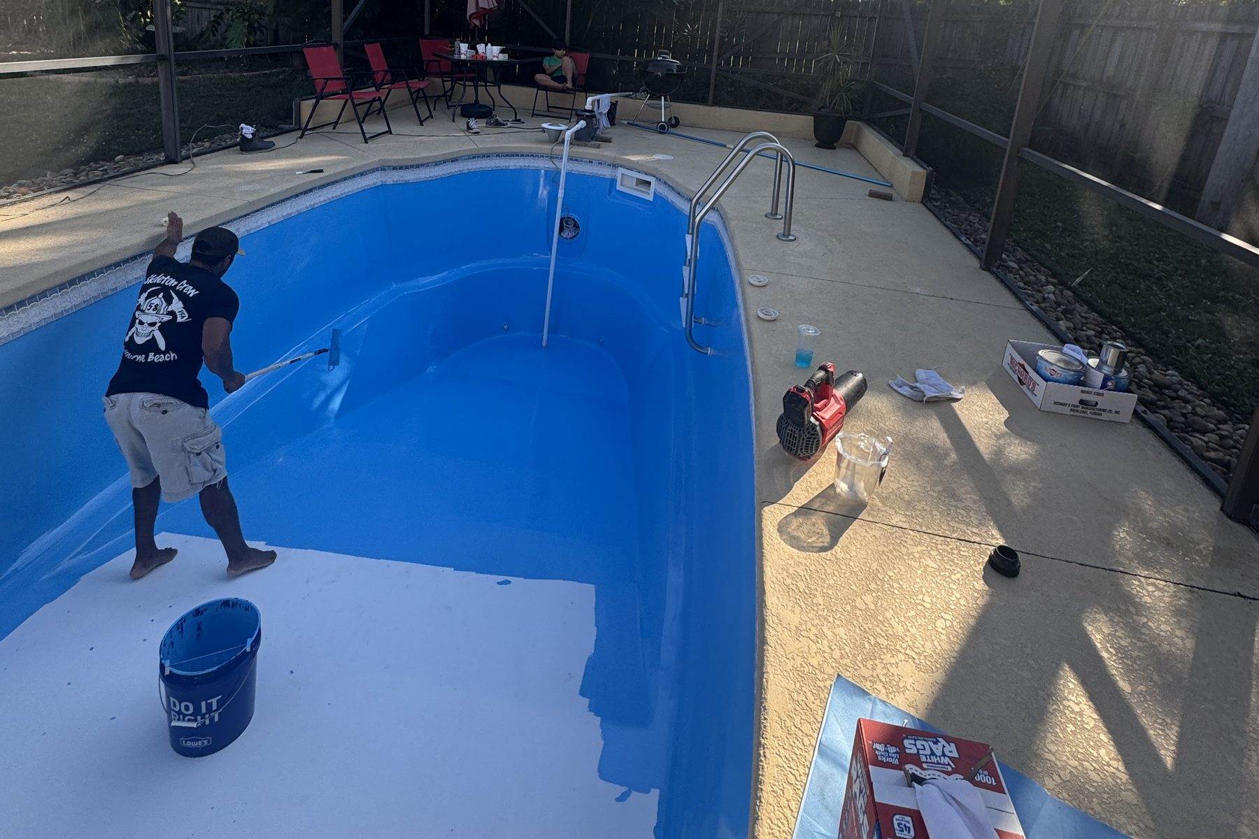 A pool interior being coated blue with a roller on an extension pole, the untreated shell still visible in the foreground