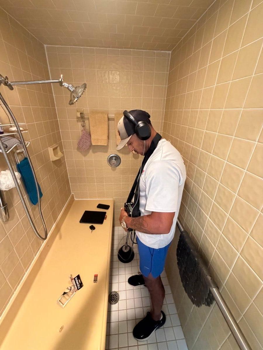 A technician wearing headphones and holding a ground microphone against a tiled shower floor, with an acoustic amplifier on a shoulder strap