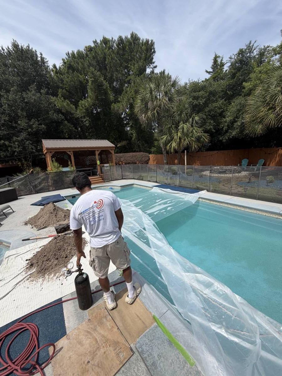 A technician at a poolside with a pressure-test cylinder and hose, the pool sheeted over, sand piles and plywood on the deck