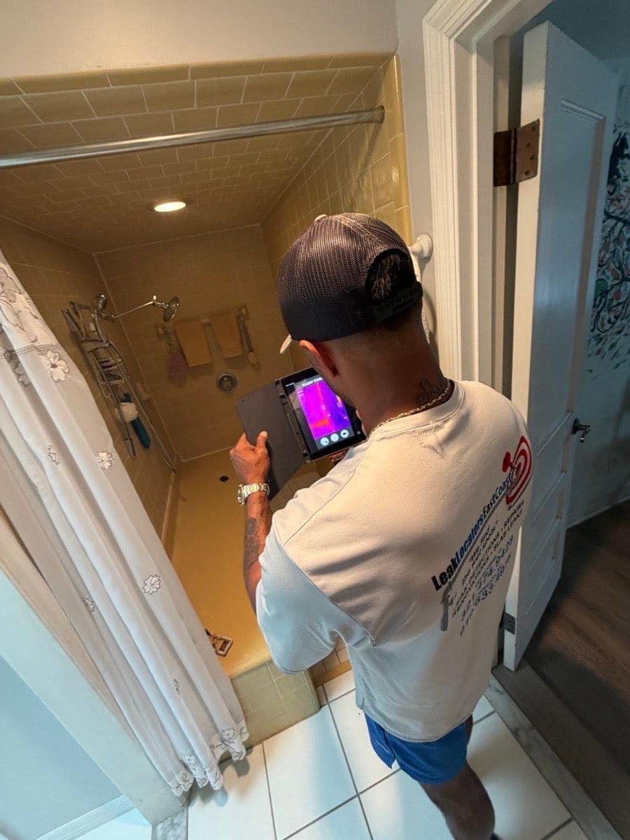 A technician holding a thermal imaging camera with the display visible, aimed at a tiled shower area