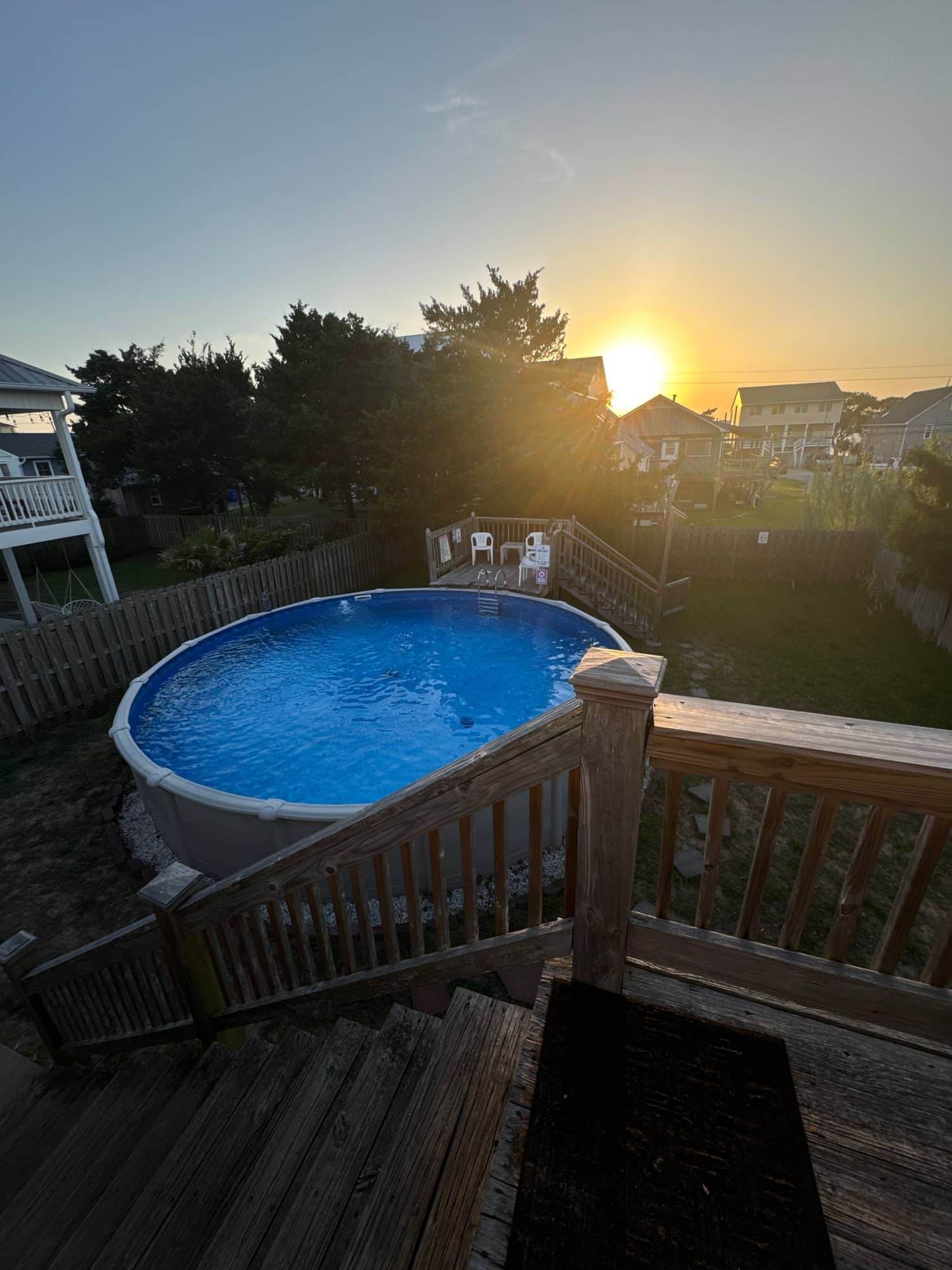 An oval above-ground pool with a vinyl liner on a residential lot at sunset, with a timber deck and steps alongside