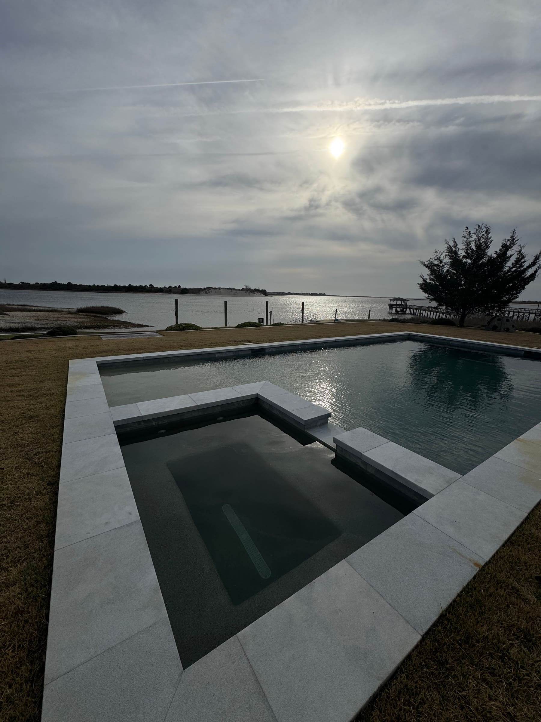 A dark-tiled pool and spa on a waterfront lawn, open water behind