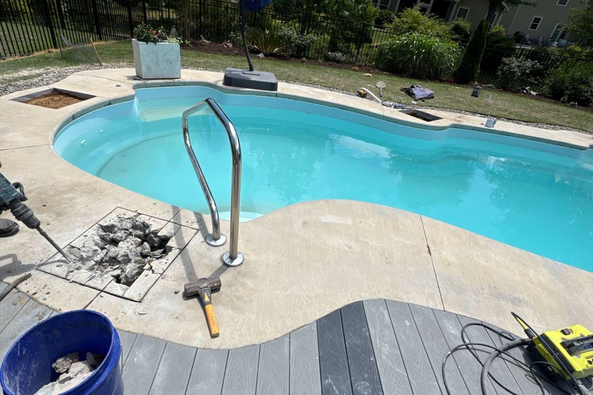 A small opened section of pool deck beside a residential pool, with broken concrete in a bucket, a sledgehammer and a jackhammer set down