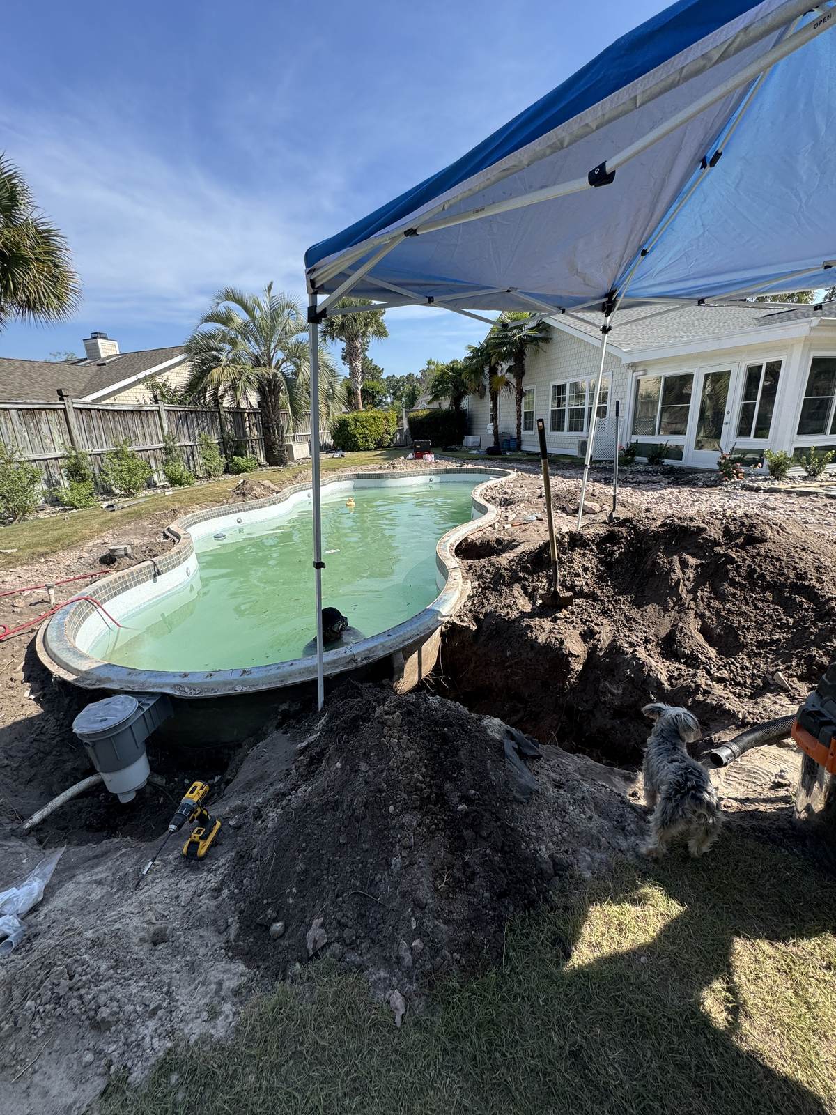 A pool with the deck edge excavated to reach the return lines, soil piled beside the opening, a work canopy over the dig and a skimmer housing in the foreground.