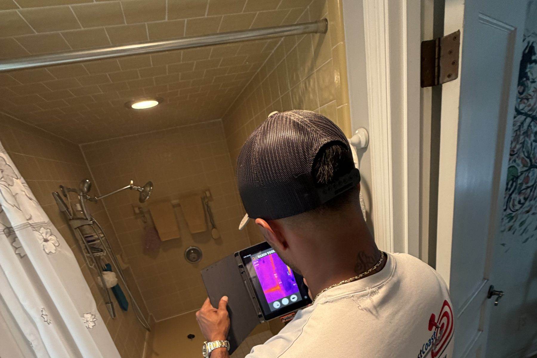 A technician holding a thermal imaging camera with the screen visible, aimed into a tiled shower